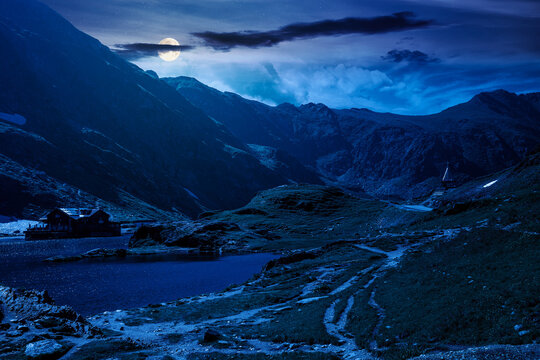 beautiful alpine scenery of fagaras mountains at night. full moon over lake in romania mountain landscape. summer weather with dark sky. rippled water near rocky slope. mysterious travel background