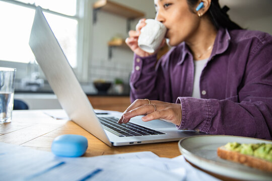 Woman working on laptop while drinking coffee in home kitchen