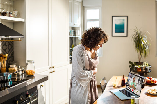 Woman with stomach pain consulting doctor online at home kitchen