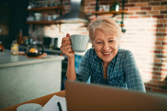 Senior woman video chatting on laptop with coffee at home kitchen