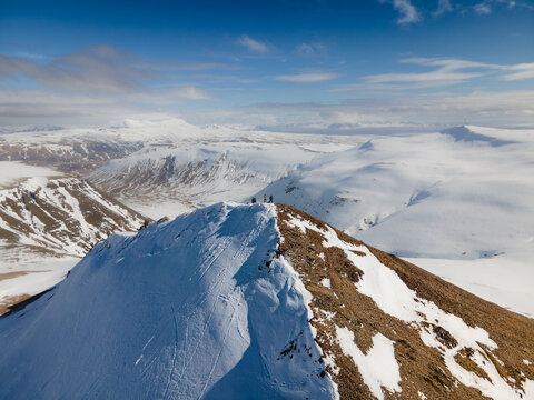 Aerial view of Moskardhshnukar mountain peak with hikers standing on the snowy summit overlooking a vast winter landscape under a blue sky Mosfellsbaer, Kjosarhreppur, Iceland.