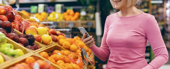 A woman stands in a grocery store looking at her phone while shopping for fruits. She is surrounded by colorful displays of apples, oranges, and other fresh produce during the afternoon hours. © Prostock-studio