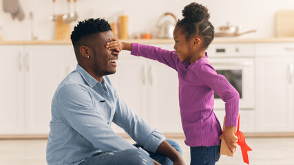 A father sits on the floor while his daughter stands in front of him covering his eyes with her hands. The kitchen is bright and has simple decor. The little girl holds a gift behind her back. © Prostock-studio