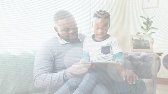 Father guiding child on couch, tilting tablet while child tapping and learning app showing screen