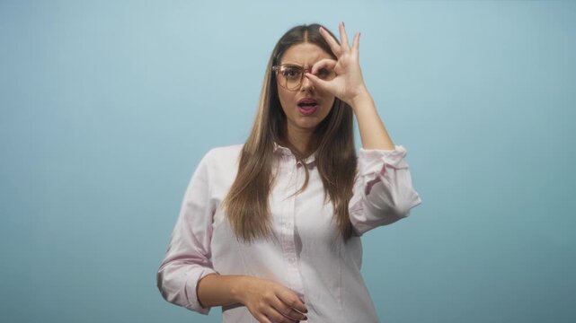 Woman wearing glasses makes ok sign over eye while peering through finger circle in studio; playful curiosity.