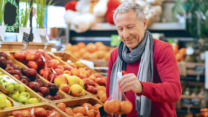A man picks out fresh oranges in a grocery store. He stands in front of a display filled with various fruits. The scene shows a busy grocery environment with many choices available. © Prostock-studio