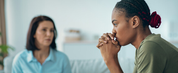 One woman is sitting with a worried expression, while another woman speaks to her. They are in a room with light colors and some plants. It looks like a discussion about feelings. © Prostock-studio