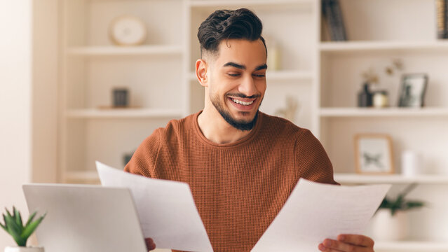 A man sits at a desk with a laptop in front of him. He holds papers in both hands and looks pleased while reading. The room has shelves and a plant nearby, suggesting a home office environment.