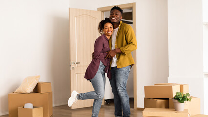 A couple stands in their new home, smiling and embracing. They are surrounded by moving boxes in a bright room. The door is open, and they look happy as they begin settling in. © Prostock-studio