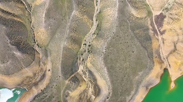 Aerial view of dry riverbeds with green water patches. Rocky slopes show layered earth tones