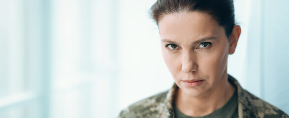 A military woman stands indoors, looking directly at the camera with a serious expression. The setting has large windows that allow light to enter. This scene takes place during the daytime. © Prostock-studio