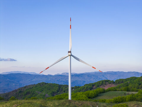 Aerial view of a large wind turbine with red-tipped blades standing on a lush green ridge overlooking the Apennine Mountains under a clear blue sky Varese Ligure, Liguria, Italy.