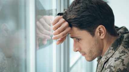 A soldier stands by a window with a look of concern on his face. He has one hand resting on his forehead. The setting is quiet, and the soldier is lost in thought. © Prostock-studio