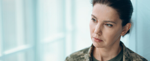 A woman in military attire stands by a large window. She gazes thoughtfully outside, lost in her thoughts. The room is bright and the atmosphere is quiet during the afternoon. © Prostock-studio