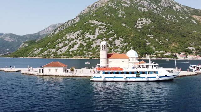 Horizontal drone video of Perast Island in Montenegro, surrounded by mountains and the sea, featuring stunning natural scenery and a tourist boat arriving at the island