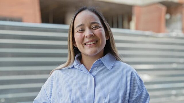 Woman smiling with visible face and blue striped shirt, tilting head toward camera in front of a building; friendly joy.