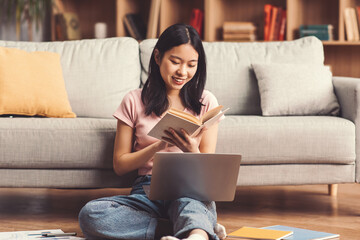 A young girl reads a book with a smile while sitting on the floor. She has a laptop open in front of her. The room has a couch and bookshelves in the background. © Prostock-studio
