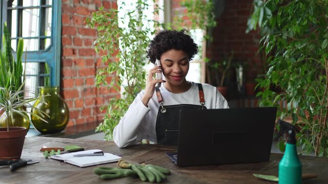 African woman florist gardener working in botanical store using laptop order plants by phone communicating with customer. Female small business owner working at flower shop. Running of own business
