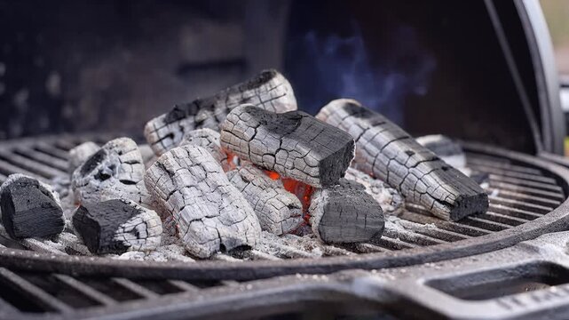 Closeup of glowing hot charcoal briquettes on a grill grate with smoke rising.
