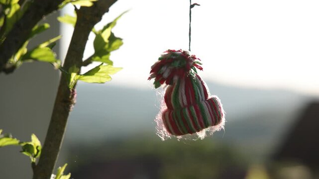 A tiny, colorful knitted cap hangs from a tree branch, glowing in the bright sunlight. The miniature beanie sways gently against a soft, blurred landscape of rolling hills.