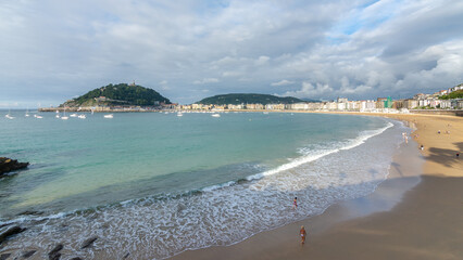 Naklejka premium People walking near the shoreline of La Concha beach with calm waves and panoramic bay view in San Sebastian, Spain.