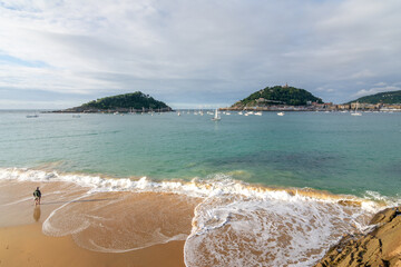 Naklejka premium Sea foam reaching the sandy shore at Ondarreta beach with panoramic coastal scenery in San Sebastian, Spain.