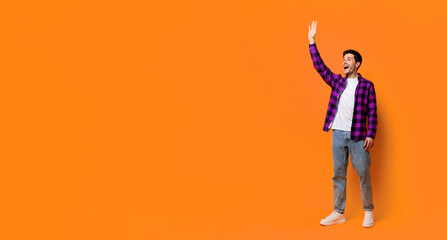 A man with dark hair wears a purple checkered shirt and jeans. He stands with a wide smile and raises his hand. The background is a bright orange wall with no distractions. © Prostock-studio