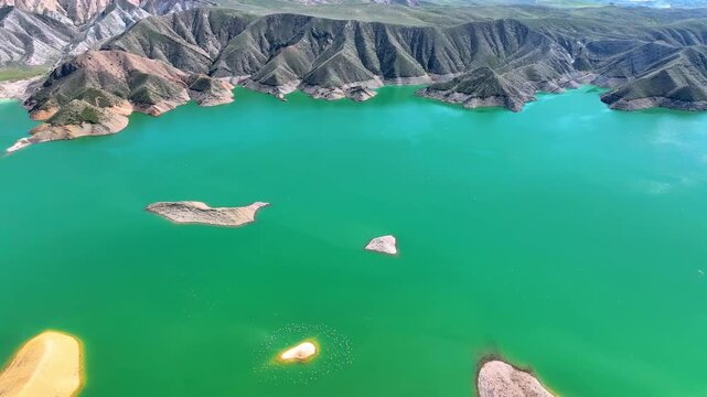 Aerial view of turquoise lake nestled between rugged brown hills. Water reflects sky, creating vivid contrast with earthy shores. Rocky outcrops and small islands dot calm surface
