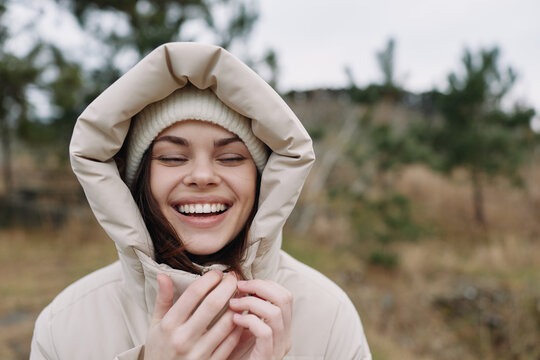 Woman with a jacket and hooded beanie outdoors, smiling in a nature landscape, showcasing casual warmth and autumn charm in a fresh outdoor setting