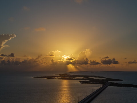 Aerial view of a long bridge stretching across the ocean towards a golden sunset with sunbeams breaking through clouds Florida, United States.