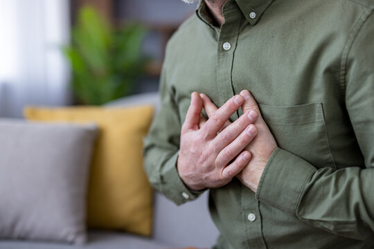 Man experiencing severe chest pain and discomfort, holding his hand over his chest while sitting on a sofa at home, indicating a potential heart attack or cardiovascular emergency