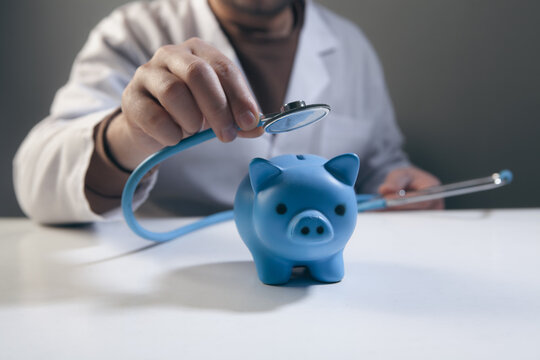 Doctor holding stethoscope with blue piggy bank on desk.