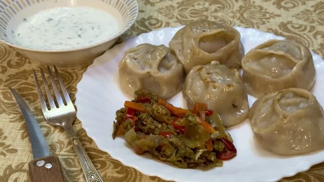 Large Manti dumplings on white plate with herbed yogurt sauce in bowl, spiced vegetable salad, fork and knife resting on patterned