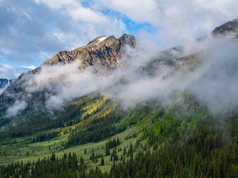 Aerial view of rugged mountain peaks shrouded in mist above a lush green valley with dense evergreen forests CO, United States.