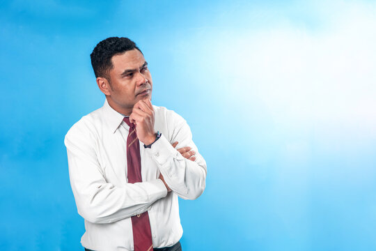 Portrait of a pensive Indonesian office worker in white shirt and tie, holding his chin while thinking deeply. Concept of problem solving and ideas.
