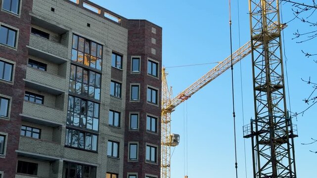 Construction crane above brick building, tower crane hoisting materials beside multistory brick shell under clear blue sky, bare