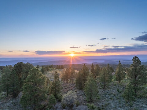 Aerial view of coniferous forest and distant canyons under a glowing sunset sky with a sunburst effect in Moab, Utah, United States.