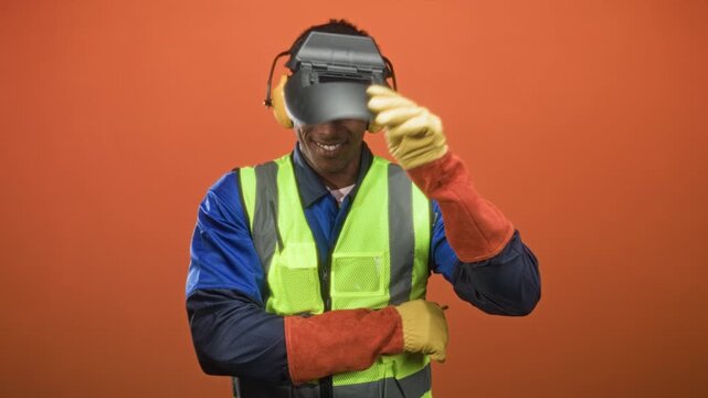 Man wearing welding helmet and reflective vest lifts helmet with gloved hand in studio, smiling face revealed; pride duty.