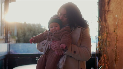 Mother holding baby in bear teddy fleece coat and beanie hat outdoors, woman in beige coat smiling gently while child raises arms during cold season afternoon together © Marco