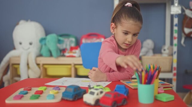 Girl drawing with pencil and holding paper at red table in preschool classroom with toy cars and stuffed octopus; calm focus learning.