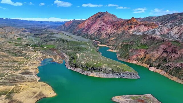 Aerial view of a winding turquoise river through rugged canyon terrain. Mountains with layered red and orange rock formations tower above. Valley floor shows green patches
