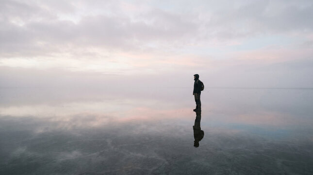 Solitary Person Standing on Reflective Shore Under Cloudy Sky