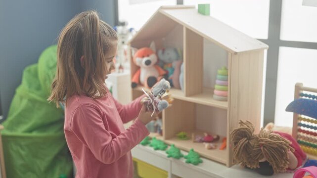 Girl holding dolls and toys with bare hands, arranging a wooden dollhouse and stacking rings on a shelf in a building; playful imagination.