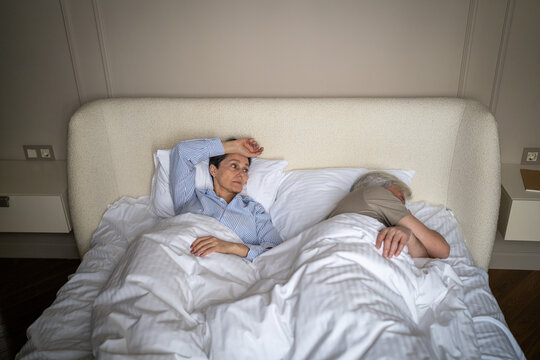 Senior married couple in serious relationship crisis, betrayed elderly wife lying in bed beside sleeping husband looking at partner with disappointment and doubt, love affair and cheating in older age