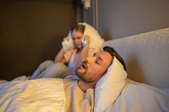Woman stressed by loud annoying sound of husband snoring, sitting on bed covering ears with pillow, exhausted female with sleep deprivation and insomnia suffering from partner snoring at night.