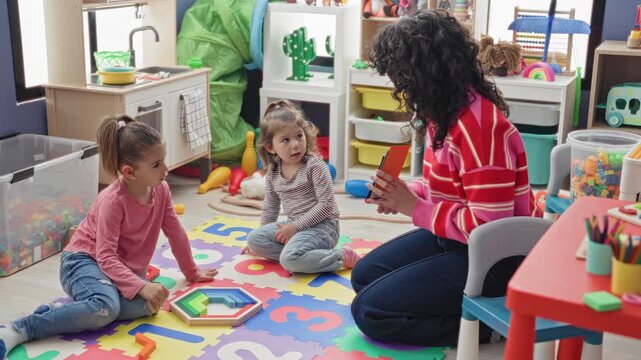 Teacher holds flashcard and points to foam mat while sitting with girls among toys in a preschool building; curiosity learning.