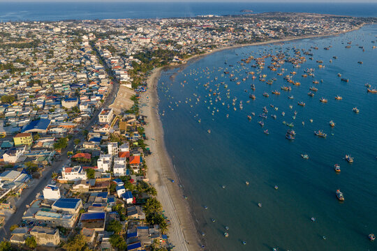 Aerial view of Mui Ne Beach featuring numerous fishing boats anchored in the turquoise bay alongside the dense coastal town buildings Phan Thiet, Binh Thuan, Vietnam.