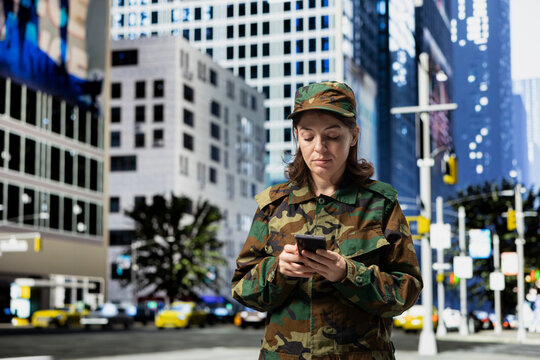 Army soldier using smartphone in city downtown to read mission debriefing details from command center. Woman wearing camouflage uniform using mobile phone to send text messages back home