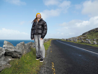 Woman stands near ocean edge, gazing at horizon. She wears puffer jacket, bright hat, and casual pants. Road winds through rocky landscape with green grass. Blue sky holds scattered clouds © mark_gusev