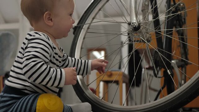 Young child investigates bicycle wheel indoors. Infant explores bicycle parts in cozy home environment. Child interacts with bicycle wheel inside house demonstrating early motor skills and curiosity